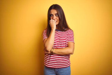 İzole sarı arka plan üzerinde duran çizgili t-shirt giyen genç güzel kadın stresli ve ağız tırnak ısırma elleri ile sinirli görünümlü. Anksiyete sorunu.