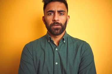 Yeşil tişörtlü genç Hintli adam izole edilmiş sarı arka plan üzerinde duruyor şüpheci ve gergin, yüzünde çapraz kollu tasvip etmeyen bir ifade. Negatif kişi.