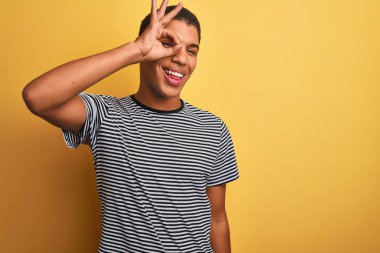 Sarı arka plan üzerinde denizci çizgili tişört giyen genç yakışıklı Arap adam el hareketleriyle gülümsüyor ve mutlu yüzüyle parmaklarının arasından bakıyor..