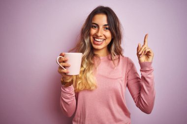 Genç ve güzel bir kadın pembe izole edilmiş arka planda kahve fincanını tutarken mutlu bir şekilde elini ve parmağını kenarı işaret ediyor.