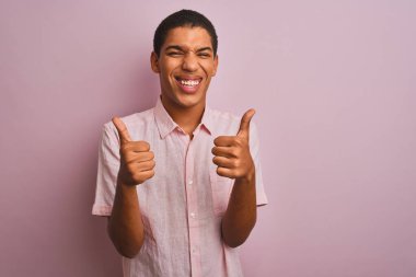 İzole edilmiş pembe arka plan başarısı tabelasının üzerinde duran rahat gömlekli genç, yakışıklı bir Arap. Elleriyle olumlu jest yapıyor. Gülümsüyor ve mutlu. Neşeli ifade ve kazanan jest.