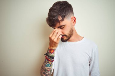 İzole edilmiş beyaz arka plan üzerinde duran dövmeli genç adam yorgun burnu ve gözleri ovuşturup yorgun ve baş ağrısı çekiyor. Stres ve hüsran kavramı.