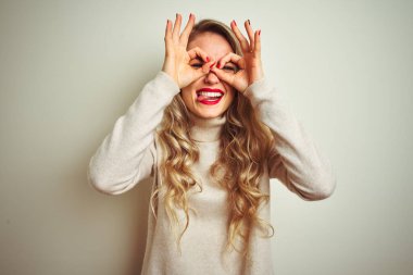 Güzel bir kadın kışın balıkçı yaka kazak giyip izole edilmiş beyaz arka plan üzerinde dürbün gibi güzel hareketler yapıyor. Dürbün dilini dışarı çıkarıyor, gözler parmaklarından bakıyor. Çılgın ifade.