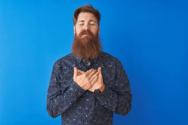 Çiçekli yaz gömleği giyen genç kızıl saçlı adam izole edilmiş mavi arka plan üzerinde duruyor elleri göğsünde, gözleri kapalı ve yüzünde minnet dolu bir jest ile gülümsüyor. Sağlık kavramı.