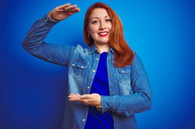Genç, kızıl saçlı, kot gömlek giyen, mavi izole edilmiş arka plan üzerinde duran, elleri büyük ve büyük beden işareti gösteren, ölçü sembolü olan bir kadın. Kameraya bakıp gülümsüyor. Ölçüm konsepti.