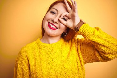 Kışlık süveter giyen genç bir kadın izole edilmiş sarı arka planda duruyor. El hareketleriyle gülümsüyor, gözleriyle mutlu yüzle parmaklarının arasından bakıyor..