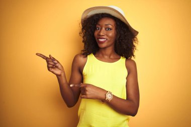 Genç Afro-Amerikalı kadın tişört ve şapka giyip izole edilmiş sarı arka planda gülümseyerek kameraya bakıyor ve iki eliyle ve parmaklarıyla yüzünü işaret ediyor..
