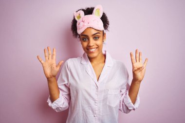 Genç Afro-Amerikalı kadın pijama giyiyor ve maske takıyor pembe arka planda 9 numarayı gösterirken kendine güvenen ve mutlu gülümseyen.