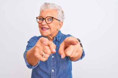 Senior grey-haired woman wearing denim shirt and glasses over isolated white background pointing to you and the camera with fingers, smiling positive and cheerful