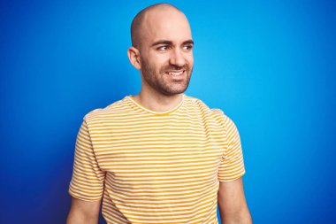 Mavi izole arka plan üzerine günlük tişört giyen sakallı genç bir adam yüzünde gülümseme, doğal bir ifade ile yan yana bakıyor. Kendinden emin gülen.