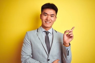 Asyalı Çinli iş adamı takım elbise ve kravat takıyor. Yüzünde kocaman bir gülümsemeyle izole edilmiş sarı arka planda duruyor. Kameraya el ve parmağıyla bakıyor..