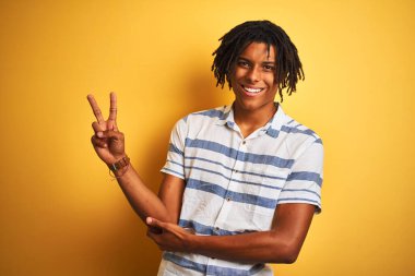 Sarı arka plan üzerinde çizgili tişörtü olan Afro-Amerikan bir adam kameraya gülümseyip zafer işareti yapıyor. İki numara..
