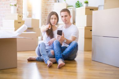 Güzel bir çift yeni evinde akıllı telefon kullanarak kamerayı işaret eden karton kutuların etrafında oturuyor. Ve sen, el işareti, ön taraftan pozitif ve kendinden emin bir hareket.