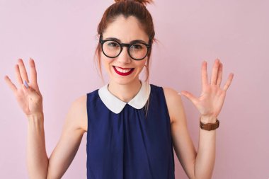 Kırmızı saçlı, atkuyruklu, zarif elbiseli ve gözlüklü, pembe arka planda, dokuz numarayı işaret eden, kendinden emin ve mutlu bir şekilde gülümseyen bir kadın..