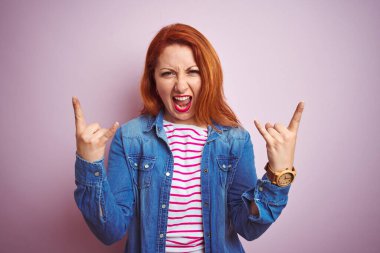 Güzel kızıl saçlı kadın kot gömlek ve çizgili tişört giyerek izole edilmiş pembe arka planda çılgın bir ifadeyle elleri havada rock sembolü yaparak bağırıyor. Müzik yıldızı. Ağır müzik konsepti.