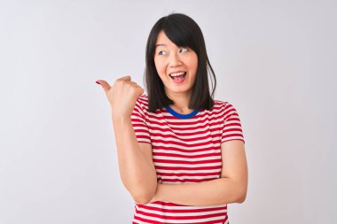 İzole edilmiş beyaz arka plan üzerinde kırmızı çizgili tişört giyen genç ve güzel Çinli bir kadın gülümsüyor ve başparmağıyla yan tarafı işaret ediyor..