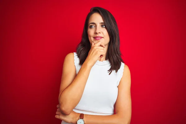 Young beautiful woman wearing white dress standing over red isolated background looking confident at the camera smiling with crossed arms and hand raised on chin. Thinking positive.