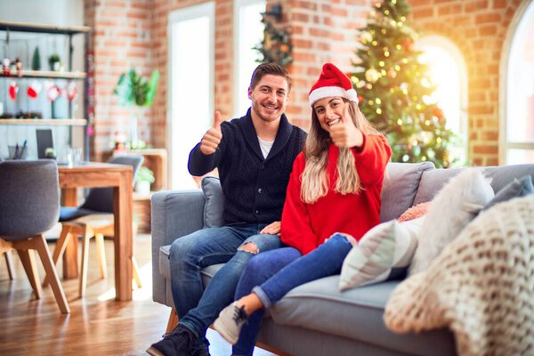 Young couple wearing santa claus hat sitting on the sofa around christmas tree at home doing happy thumbs up gesture with hand. Approving expression looking at the camera with showing success.