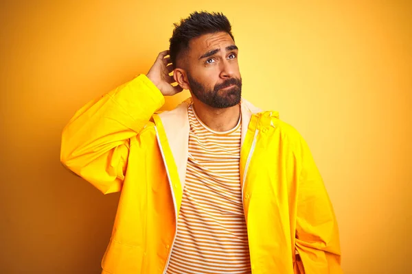 Young indian man wearing raincoat standing over isolated yellow background confuse and wonder about question. Uncertain with doubt, thinking with hand on head. Pensive concept.