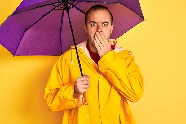 Young man wearing raincoat holding purple umbrella standing over isolated yellow background cover mouth with hand shocked with shame for mistake, expression of fear, scared in silence, secret concept