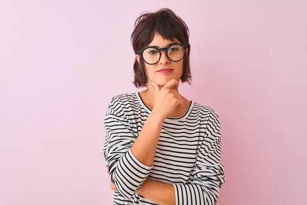 独立したピンクの背景にストライプのTシャツとメガネを身に着けている若い美しい女性は 交差腕と手で笑顔でカメラを見て自信を持ってあごに提起 肯定的な考え — ストック写真