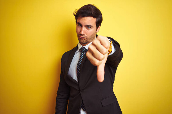 Young handsome businessman wearing suit and tie standing over isolated yellow background looking unhappy and angry showing rejection and negative with thumbs down gesture. Bad expression.