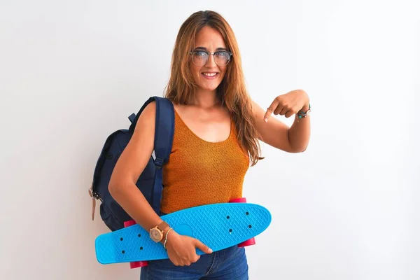 年轻的红头发女学生 背着背包和滑板 背景偏僻 手指手画脚 非常高兴 — 图库照片