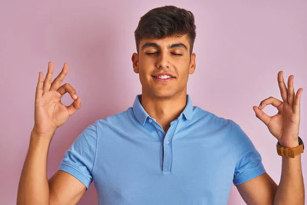 穿着蓝色马球站在孤立的粉色背景上的印度年轻人放松 闭上双眼微笑 用手指做冥想手势 瑜伽概念 — 图库照片