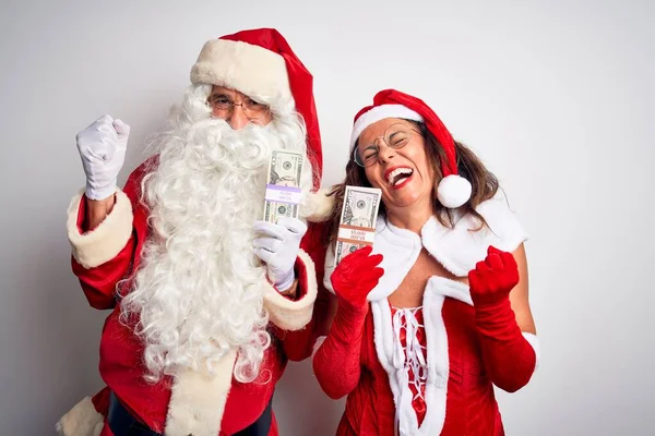 Senior couple wearing Santa Claus costume holding dollars over isolated white background very happy and excited doing winner gesture with arms raised, smiling and screaming for success. Celebration concept.