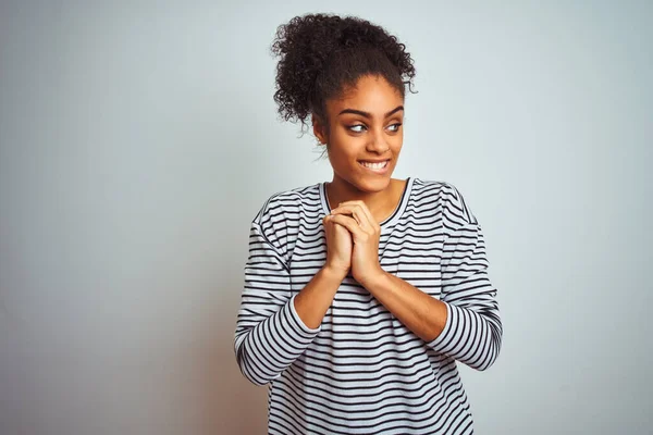 African american woman wearing navy striped t-shirt standing over isolated white background laughing nervous and excited with hands on chin looking to the side