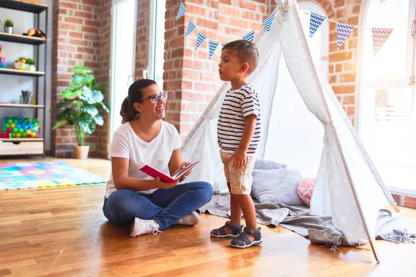 Beautiful teacher and toddler boy sitting on the floor playing inside  tipi at kindergarten