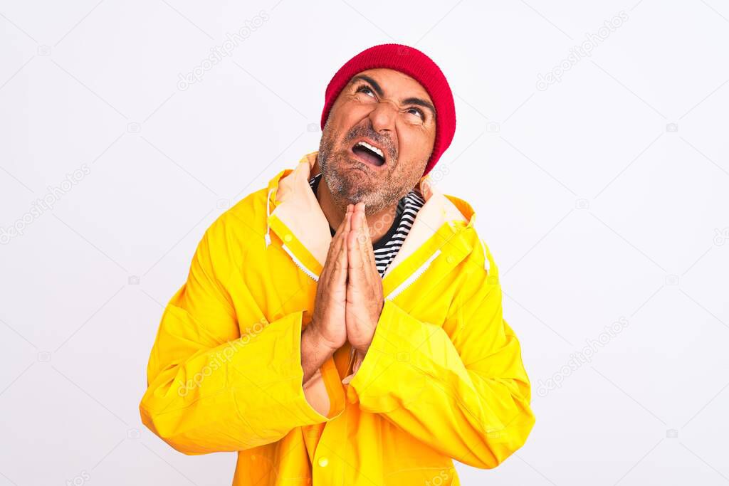Middle age man wearing rain coat and woolen hat standing over isolated white background begging and praying with hands together with hope expression on face very emotional and worried. Asking for forgiveness. Religion concept.