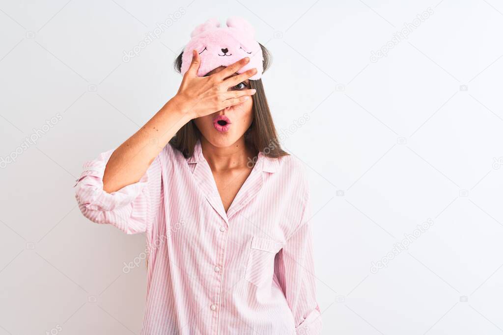 Young beautiful woman wearing sleep mask and pajama over isolated white background peeking in shock covering face and eyes with hand, looking through fingers with embarrassed expression.