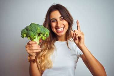 Gri izole arka planda brokoli yiyen genç ve güzel bir kadın bir fikir ya da soru işareti ile şaşırdı.