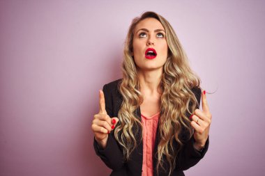 Zarif bir ceket giyen genç ve güzel bir iş kadını. Pembe izole edilmiş arka planda duruyor. Şaşırmış ve şaşırmış..