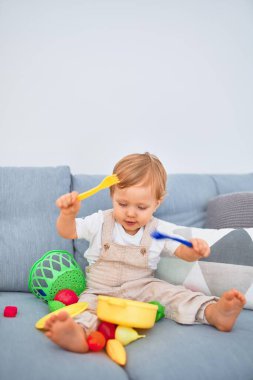 Sevimli sarışın çocuk kanepede oturup evde plastik yemek oyuncaklarıyla oynuyor.