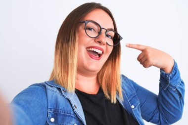 Kot gömlek ve gözlük takan güzel bir kadın izole edilmiş beyaz arka plan üzerinde selfie yapar. Elle ve parmakla göstererek mutlu olur.