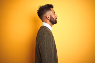 Yeşil kazak ve tişört giyen genç Hintli adam, yalıtılmış sarı arka plan üzerinde duruyor, yan yana bakıyor, rahat profil pozu, kendine güvenen bir gülümsemeyle doğal bir yüz..