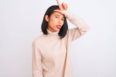 Young chinese woman wearing turtleneck sweater standing over isolated white background making fun of people with fingers on forehead doing loser gesture mocking and insulting.