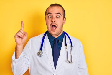 Paltolu ve steteskoplu genç doktor izole edilmiş sarı arka planda duruyor ve başarılı bir fikirle işaret ediyor. Heyecanlı ve mutlu. Bir numara..