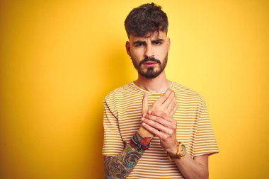 İzole edilmiş sarı arka plan üzerinde duran çizgili tişörtlü genç bir adam. Ellerinde ve parmaklarında ağrı, iltihaplanma.