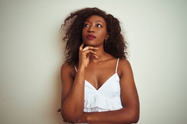 Genç afro-amerikan kadın üzerinde izole beyaz arka plan üzerinde duran t-shirt giyen çene üzerinde el soru, dalgın ifade düşünme. Düşünceli bir yüzle gülümsüyor. Şüphe kavramı.