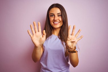 İzole edilmiş pembe arka plan üzerinde duran rahat tişört giyen genç ve güzel bir kadın dokuz numaralı parmaklarıyla işaret ederken kendinden emin ve mutlu bir şekilde gülümsüyor..