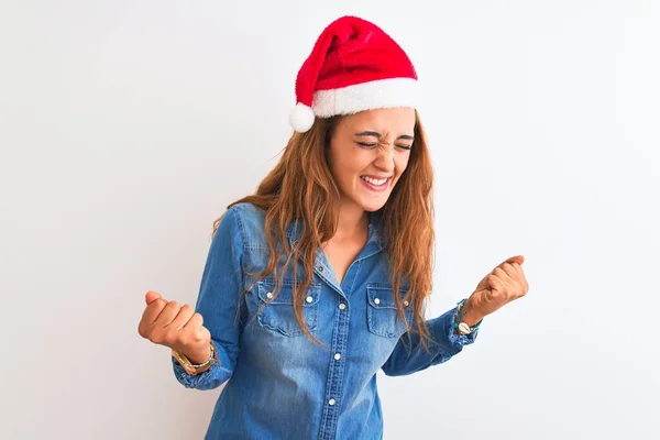 孤立した背景の上にクリスマスの帽子を身に着けている若い美しい赤毛の女性は非常に満足し 腕を上げ 笑顔と成功のために叫んで勝者のジェスチャーを行うことに興奮しています お祝いのコンセプト — ストック写真
