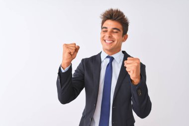 İzole edilmiş beyaz arka plan üzerinde duran takım elbiseli genç yakışıklı işadamı kolları havada, gülümseyen ve başarı için bağıran mutlu ve heyecanlı bir jest yapıyor. Kutlama konsepti.