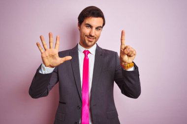 Genç yakışıklı iş adamı takım elbise ve kravat takıyor pembe arka plan üzerinde duruyor ve altı numaralı parmaklarıyla işaret ederken kendinden emin ve mutlu gülümsüyor..