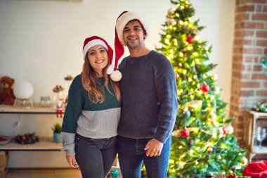 Mutlu ve kendinden emin gülümseyen genç bir çift. Evde Noel Baba şapkası takmış Noel ağacının etrafında dikiliyor.