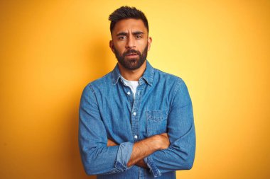 İzole edilmiş sarı arka plan üzerinde duran kot gömlekli genç Hintli adam şüpheci ve gergin, yüzünde çapraz kollu tasvip etmeyen ifade. Negatif kişi.