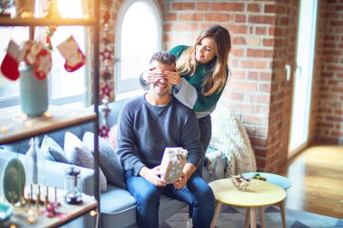 Mutlu ve kendinden emin gülümseyen genç bir çift. Erkeğin gözlerini kapayan bir kadın Noel süslemeleriyle süpriz yapıyor.