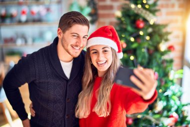 Mutlu ve kendinden emin gülümseyen genç bir çift. Ayakta durmak ve sarılmak, evdeki Noel ağacının etrafında kamerayla selfie çektirir.